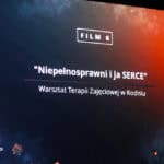 Widok na ekran, na którym wyświetlony jest slajd z napisem: „Film 6. Niepełnosprawni i ja SERCE. Warsztat Terapii Zajęciowej w Kodniu”. Na ekranie dominują barwy niebieskie i czerwone. Kolory przenikają się, tworząc gradient. W lewym górnym rogu logo Caritas Diecezji Siedleckiej. W lewym dolnym logo Festiwalu Filmowego Twórczości Osób Niepełnosprawnych. W prawym górnym rogu logo Warsztatu Terapii Zajęciowej w Białej Podlaskiej. W lewym dolnym rogu logo XII Edycji – w kształcie klatki kliszy filmowej