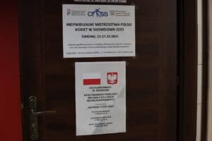 Dwa plakaty informacyjne przymocowane do drewnianych drzwi. Górny plakat ogłasza: "INDYWIDUALNE MISTRZOSTWA POLSKI KOBIET W SHOWDOWN 2025. ŚWIDNIK, 23-27.10.2025" i informuje o współfinansowaniu z Ministerstwa Sportu i Turystyki oraz PFRON, z organizacją Stowarzyszenia "CROSS". Dolny plakat z symbolami Polski (flaga i godło) informuje o dofinansowaniu ze środków Państwowego Funduszu Rehabilitacji Osób Niepełnosprawnych, zadanie "WSPÓLNY START 2025", z kwotami dofinansowania i całkowitej wartości projektu