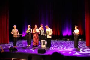 Grupa siedmiu osób, stoi na scenie w końcowej fazie wydarzenia, podczas wręczania nagród i dyplomów. Niektóre osoby trzymają w rękach dyplomy, a w centrum widoczny jest karton z nagrodą (głośnikiem). Scena jest oświetlona fioletowym światłem na tle purpurowej kurtyny, a na podłodze widoczny jest jasny, wirujący wzór