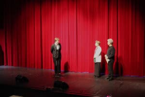 Scena teatralna z ciężką, czerwoną kurtyną w tle. Na scenie stoją trzy osoby: kobieta w czarnym stroju (Joanna Olszewska), mówi do mikrofonu, patrzy w kierunku stojących obok: kobiety w białej marynarce – Karoliny Czajki i mężczyzny w czarnym stroju - Michała Stanowskiego