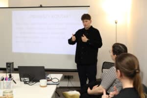 Scena ze szkolenia: prowadząca (Wioletta Stępniak) w czarnej koszuli stoi przodem do sali, gestykulując obydwoma kciukami w górę. Za nią wyświetlany jest slajd z nagłówkiem "PIERWSZY KONTAKT" i listą podstawowych zwrotów, takich jak: dzień dobry, dobry wieczór, do widzenia, przepraszam, itd.