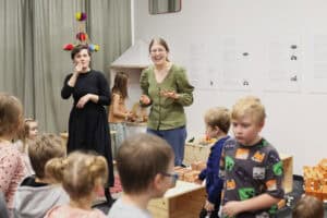 Otwarcie wernisażu wystawy. Od lewej tłumaczka języka migowego, z prawej roześmiana kobieta w okularach. Pierwsza, w czarnej sukience trzyma palec przy ustach, a druga w zielonej bluzce opowiadając, gestykuluje. Dzieci stoją w półkolu, słuchają. W tle widać minimalistyczną scenografię: domek z białym dachem i sensoryczną wiszącą instalację z kolorowymi pomponami