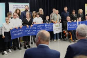 Grupa osób stoi na scenie. Osoby te trzymają niebieskie tablice z białymi napisami, m.in.: „OTULINKA ZAMOŚĆ", „KOMPLEKS SPORTOWY ZAMOŚĆ", „RODZINNY DOM ZAMOŚĆ", „CAS 3 Mieszkanie Treningowe w Biłgoraju" oraz „CASON BIAŁOBRZEGI". Kilka osób siedzi na wózkach. Na pierwszym planie widoczne są tyły głów dwóch mężczyzn w garniturach, którzy patrzą w kierunku sceny. W tle, na ścianie, widoczny jest fragment banera z napisem „STOWARZYSZENIE POMOCY DZIECIOM NIEPEŁNOSPRAWNYM"