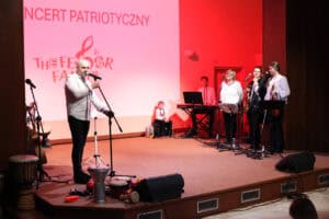 Mężczyzna (Marek Fedor) w białej koszuli stoi na scenie oświetlonej na czerwono i przemawia do mikrofonu. W tle widoczny jest ekran z napisem „KONCERT PATRIOTYCZNY" oraz część zespołu muzycznego, w tym kobieta i młodzi ludzie, z instrumentami i keyboardem. Na pierwszym planie, na podłodze sceny, leżą instrumenty perkusyjne