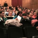 Sala kinowa z rzędami czarnych foteli wypełniona publicznością; widzowie, w większości osoby dorosłe siedzą, trzymają w rękach ulotki lub notatki; na pierwszym planie po prawej stronie widać kobietę w beżowym kombinezonie schyloną w kierunku jednej z kobiet siedzących na widowni