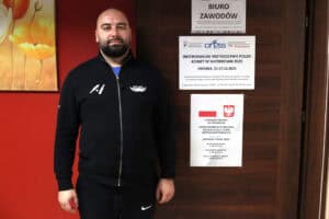 Mężczyzna z brodą (Sebastian Michailidis), ubrany w czarny rozpinany dres z białym logo, stoi na wprost obiektywu, uśmiechając się lekko. Stoi na tle czerwonej ściany i ciemnych, drewnianych drzwi. Na drzwiach zawieszone są plakaty informacyjne dotyczące "Indywidualnych Mistrzostw Polski Kobiet w Showdown 2025" oraz dofinansowania PFRON w ramach programu "WSPÓLNY START 2025"