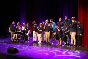 Na scenie stoi duża grupa młodych osób z zespołu "Rozśpiewany Zakątek" w Puławach, w większości ubranych w ciemne koszulki z logo oraz jasne spodnie. W grupie widoczna jest jedna osoba na wózku inwalidzkim. Uczestnicy są zwróceni do widowni. Scena oświetlona jest fioletowym światłem na tle purpurowej kurtyny