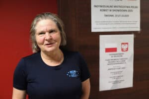 Średnie zbliżenie uśmiechniętej kobiety (Joanna Pożarycka) w granatowej koszulce z małym logo "OKS WIM Olsztyn". Kobieta ma krótkie włosy i jest oparta o ścianę. Po prawej, w tle, na drewnianych drzwiach, widoczne są dwa plakaty informacyjne o "Indywidualnych Mistrzostwach Polski Kobiet w Showdown 2025" oraz o dofinansowaniu z PFRON