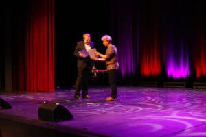 Uroczystość wręczenia dyplomów: dwóch mężczyzn stoi na środku sceny. Michał Stanowski (po prawej) wręcza dyplom Mateuszowi Dudkowi (po lewej). Mateusz ma na sobie elegancki ciemny garnitur, a Michał brązową koszulę. Scena jest oświetlona fioletowym światłem, a w tle widać kurtyny podświetlone na czerwono i fioletowo