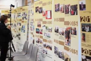 Ściana z banerami prezentującymi historię Stowarzyszenia „Krok za Krokiem" w ujęciu chronologicznym od 1990 roku. Banery w kolorze beżowym i brązowym zawierają daty, krótkie opisy i małe zdjęcia ilustrujące wydarzenia z tych lat. Po lewej stronie, obok statywu z kamerą stoi kobieta w czarnym stroju, patrzy na banery. Na pierwszym planie widoczne są trzy szare, minimalistyczne krzesła