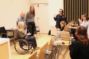 Na pierwszym planie po lewej na wózku siedzi Angelika Chrapkiewicz-Gądek. Ma jasne włosy, klaszcze w dłonie. Nad nią stoją dwaj mężczyźni (Maciej Książko i Piotr Kękuś) – jeden w koszuli w kratę i młodszy z długimi włosami – patrzą w stronę kobiety. Po prawej, kobieta (Anna Bieganowska-Skóra) w okularach i ciemnym żakiecie, stoi za stołem, uśmiecha się szeroko i gestykuluje. Za stołami widać rzędy słuchaczy, niektórzy z nich klaszczą w dłonie. Na pierwszym planie po prawej, na tle widowni, widoczna jest kobieta robiąca zdjęcie aparatem