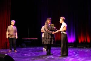 Uroczystość wręczenia dyplomów: Małgorzata Pietralska w ciemnej, wzorzystej sukience z falbanami trzyma czarny dyplom i ściska dłoń Karoliny Czajki w białej koszulce i szerokich czarnych spodniach. Po lewej stronie uśmiecha się mężczyzna w brązowej etnicznej koszuli (Michał Stanowski). Scena jest oświetlona fioletowo-czerwonym światłem