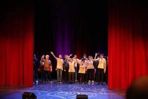 Duża grupa uczestników wydarzenia stoi na scenie między zamykającą się czerwoną kurtyną i macha do widowni na pożegnanie. Osoby są ubrane w jasne T-shirty i mają podniesione ręce. W tle widoczna fioletowa kurtyna, a na podłodze wije się okrągły świetlisty wzór