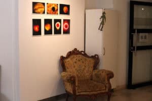 Fragment wnętrza galerii z jasnymi ścianami i podłogą wyłożoną beżowymi płytkami. Na ścianie w centralnej części wiszą dwa rzędy małych, oprawionych zdjęć, prezentujących abstrakcyjne, świetliste kształty w odcieniach czerwieni, pomarańczu i żółci. Na pierwszym planie stoi rzeźbiony, tapicerowany fotel w stylu barokowym, obity wzorzystą, musztardową tkaniną. Po prawej stronie widać białą szafę z zawieszoną na niej zieloną figurką i przeszklone drzwi