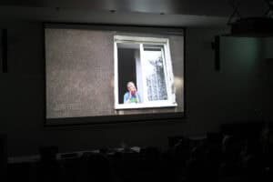 Projekcja filmu w ciemnej sali, pogrążonej w ciemności. Na ekranie wyświetlony jest kadr przedstawiający fasadę budynku o szarej, chropowatej ścianie. Centralnie umieszczone jest prostokątne, białe okno z częściowo otwartym skrzydłem. W otwartym oknie widać uśmiechniętego mężczyznę (Piotr Kękuś) w niebieskiej koszulce, który opiera się na ramie okiennej.