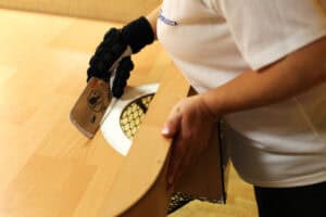 Zbliżone ujęcie dłoni zawodniczki w białej koszulce i czarnej rękawicy z ochraniaczami. Dłoń trzyma drewnianą rakietkę w kształcie prostokąta ze ściętymi rogami i uderza nią w kierunku okrągłego otworu bramkowego, zakrytego siatką