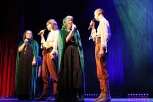 Występ muzyczny ŚDS Akademia Artystyczna: cztery osoby w kostiumach na scenie, śpiewają do mikrofonów. Dwie kobiety mają długie, zielone peleryny z kapturami w stylu fantasy. Dwie inne osoby mają na sobie luźne, jasne koszule i brązowe spodnie. Scena jest oświetlona mocnymi, niebieskimi i zielonymi światłami