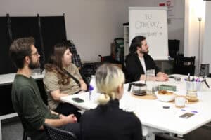 Ujęcie z boku, przedstawiające fragment grupy uczestników szkolenia siedzących przy białym stole. Uczestnicy, w tym brodaty mężczyzna w zielonym swetrze i okularach, aktywnie słuchają (lub obserwują miganie) prowadzącej, która znajduje się poza kadrem. Na stole widać notatniki i napoje