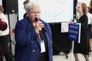 Zbliżenie na kobietę (Maria Król) w niebieskim, tweedowym żakiecie, okularach i z krótkimi, jasnymi włosami, która mówi do mikrofonu. Ma ona zawieszoną na szyi smycz z identyfikatorem. Tło jest rozmyte, ale po prawej stronie widoczna jest młoda kobieta trzymająca niebieską tabliczkę z napisem „KOMPLEKS SPORTOWY ZAMOŚĆ". Za kobietą przemawiającą widoczny jest duży ekran z grafiką