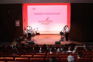 Szeroki plan sceny z widownią. Na scenie oświetlonej czerwienią, znajduje się zespół występujący podczas „Koncertu Patriotycznego”. Z lewej mężczyzna przy mikrofonie, za nim inni muzycy. W tle duży ekran z napisem „KONCERT PATRIOTYCZNY" i logo „The Fedor Family". Na pierwszym planie widoczne są pomarańczowe fotele z publicznością oglądającą występ. Po lewej stronie sceny, na ścianie wisi krzyż i godło Polski.