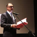 Mężczyzna (Jakub Semeniuk) w ciemnym garniturze i okularach stoi na scenie i przemawia do mikrofonu, trzymając w rękach otwartą, czerwoną teczkę z dokumentami; za nim na ekranie wyświetla się logo Caritas Diecezji Siedleckiej