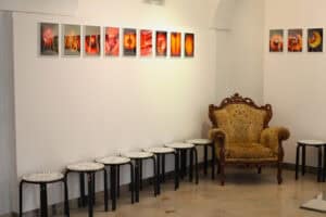 Jasna sala wystawowa z białymi ścianami. Na lewej ścianie wzdłuż góry wisi rząd dziewięciu prostokątnych fotografii, przedstawiających abstrakcyjne formy o ciepłych, pomarańczowo-czerwonych barwach, wyglądających jak podświetlone wnętrza owoców. Poniżej, na podłodze, stoi siedem czarnych, metalowych taboretów z białymi, okrągłymi siedziskami z napisami, ustawionych w rzędzie. Przy prawej ścianie znajduje się zdobiony, barokowy fotel obity beżową, wzorzystą tkaniną