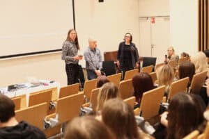 Sala wykładowa. Trzy osoby (Maciej Książko, Piotr Kękuś, Anna Bieganowska-Skóra) stoją przodem do widowni przy ekranie projekcyjnym. Kobieta po prawej, ubrana w ciemny żakiet, ma poważny wyraz twarzy i patrzy na publiczność. Na dalszym planie, po prawej, siedzi młoda kobieta, robi zdjęcie telefonem. Na pierwszym planie widoczne są tyły głów słuchaczy siedzących w rzędach krzeseł z jasnego drewna.