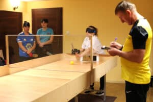 Scena z meczu showdown. Zawodniczka (Teresa Janakowska) w białej koszulce i czarnej opasce na oczach, zasłaniającej wzrok, uderza rakietką piłkę, która toczy się po drewnianym stole w kierunku aparatu. Stół ma bandy boczne i przezroczystą, środkową przegrodę. Po prawej sędzia w żółtej koszulce zapisuje wynik na notesie. Dwie osoby w opaskach zsuniętych na czoło, stoją z tyłu za stołem, obserwując grę