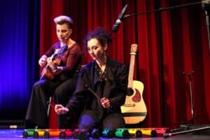 Dwie kobiety podczas występu muzycznego na scenie z czerwoną kurtyną w tle. Na pierwszym planie kobieta (Dorota Skałecka) ubrana w czarną kurtkę, siedzi na scenie i przy pomocy pałeczek gra na kolorowych tubach perkusyjnych. Kobieta z tyłu (Karolina Czajka) gra na gitarze,. W tle stoi dodatkowa gitara akustyczna.