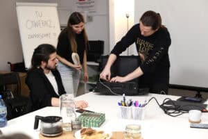 Interaktywna część szkolenia: dwóch mężczyzn i jedna kobieta pochylają się nad otwartą czarną, profesjonalną walizką transportową ze sprzętem. Mężczyzna w czarnej koszuli dotyka kabli wewnątrz walizki, demonstrując lub przygotowując sprzęt. W tle tablica z napisem "TU OTWIERACZ, ZAPRASZAMY"