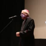 Mężczyzna (biskup siedlecki ks. Kazimierz Gurda) ubrany w czarną sutannę z czerwonymi akcentami, stoi na scenie i przemawia do mikrofonu, a w tle na ekranie wyświetla się białe logo Warsztatu Terapii Zajęciowej w Białej Podlaskiej