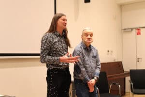 Dwóch mężczyzn (Maciej Książko i Piotr Kękuś) stoi obok siebie na tle białego ekranu projekcyjnego i jasnej ściany. Od lewej mężczyzna (Maciej Książko) z długimi, ciemnymi włosami w koszuli we wzory i czarnych spodniach z ozdobnymi sznurowaniami po bokach. Ma otwarte dłonie i wygląda, jakby mówił do kogoś spoza kadru. Po prawej, obok niego, mężczyzna w koszuli w kratę i dżinsach, ze skrzyżowanymi ramionami na wysokości pasa. Tło jest jasne, widać też fragment ciemnego pianina z prawej strony.