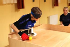 Zbliżenie na zawodniczkę (Ewa Ćwiek) podczas meczu showdown. Kobieta w niebiesko-czarnej koszulce i dużej, niebiesko-czarnej opasce zasłaniającej oczy, jest pochylona nad narożnikiem drewnianego stołu. Ma na prawej dłoni czerwoną rękawicę i trzyma rakietkę tuż przy żółtej piłce, którą zamierza uderzyć w kierunku bramki. W prawym górnym rogu, niewyraźnie, widać siedzącą obserwatorkę w okularach