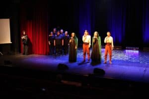 Występ ŚDS "Akademia Artystyczna". Szerokie ujęcie sceny teatralnej podczas występu. W centralnej części cztery osoby w kostiumach fantasy (dwie w zielonych płaszczach i dwie w jasnych koszulach), śpiewają do mikrofonów. Po lewej stronie ekran projekcyjny i kobieta w czarnym stroju (tłumaczka języka migowego). W tle stoi grupa chórzystów. Scena jest oświetlona światłami w kolorze niebieskim