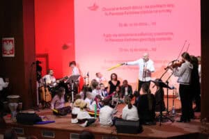 Scena oświetlona na biało-czerwono. Centralna postać, mężczyzna w białej koszuli, gestykuluje i dyryguje, stojąc obok dzieci siedzących na podłodze. Po prawej stronie widać młodą kobietę grającą na skrzypcach. Na dużym ekranie w tle wyświetlany jest tekst piosenki, zawierający frazy "Oj da, oj da dana...A gdy się szczęśliwie zakończy powstanie, to Pierwsza Kadrowa gwardyją zostanie”