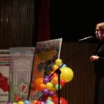 Kobieta (Justyna Gorczyca) w czarnym stroju stoi na scenie po prawej stronie i przemawia do mikrofonu, oświetlona silnym światłem bocznym, a po jej lewej stronie widoczne są plakaty reklamowe, w tym jeden promujący Festiwal Filmów Twórczości Osób Niepełnosprawnych oraz dekoracja z kolorowych balonów