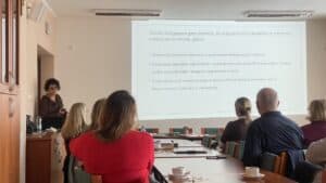 Sala wykładowa; uczestnicy siedzą przy stolikach, wpatrują się w slajd wyświetlony na ścianie. Przedstawia on charakterystykę modelu Seligmana w kontekście wielowarstwowości seksualności. Po lewej stronie przed uczestnikami stoi prowadząca i wykłada temat