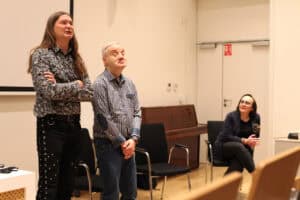 Dwóch mężczyzn (Maciej Książko i Piotr Kękuś) stoi na pierwszym planie, patrzą w górę lub na publiczność poza kadrem. Mężczyzna po lewej, z długimi włosami i w koszuli we wzory, ma skrzyżowane ramiona. Obok mężczyzna w koszuli w kratę, stoi z dłońmi splecionymi na wysokości pasa. W tle, po prawej stronie, siedzi na krześle kobieta (Anna Bieganowska-Skóra) w okularach i ciemnym żakiecie. Spogląda w górę z delikatnym uśmiechem.