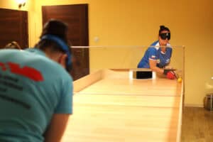 Ujęcie podczas meczu showdown z perspektywy jednego z graczy. Na pierwszym planie plecy zawodnika w niebieskiej koszulce. Po drugiej stronie drewnianego stołu, przedzielonego przezroczystą pleksiglasową przegrodą, widoczna jest przeciwniczka w niebieskiej koszulce i czarnej opasce na oczach, trzymająca rakietkę i przygotowująca się do uderzenia żółtej piłki w kierunku bramki