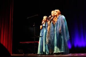 Występ tria "Mlekołaki". Trzy młode kobiety stoją obok siebie na scenie i śpiewają do mikrofonów. Ubrane są w jasnoniebieskie, zwiewne suknie i przezroczyste, błękitne płaszcze, z wiankami lub opaskami na głowach. Tło jest ciemne, z fragmentem czerwonej kurtyny po lewej i niebieskim światłem po prawej