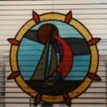 W oknie z żaluzjami wisi kolorowy, szklany witraż z motywem morskim