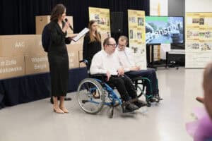 Na pierwszym planie po lewej stoi wysoka kobieta w długiej, ciemnej sukience, która trzyma mikrofon i kartkę z notatkami. Obok niej stoi druga kobieta oraz dwóch mężczyzn siedzących na wózkach inwalidzkich, ubranych w białe koszule. Za nimi po lewej widać rząd dużych, tekturowych pudeł z napisami m.in. „dr Zofia Kułakowska" oraz „Wsparcie Belgia". Po prawej znajduje się duży monitor z napisem "Transport" oraz banery z chronologią wydarzeń stowarzyszenia