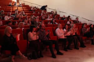 Publiczność siedzi na pomarańczowych fotelach w sali koncertowej, oświetlonej ciepłym światłem. Wszyscy widoczni na pierwszym i dalszych planach biją brawo. Na pierwszym planie widać kilka osób, w tym mężczyznę robiącego zdjęcie lub nagrywającego telefonem, oraz kobietę trzymającą małą polską flagę.