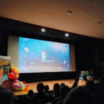 Szerokie ujęcie sali kinowej; na dużym ekranie wyświetla się plansza z napisem "XII Edycja" i hasłem "Niepełnosprawni i Ja" na tle kwiatów, widziana z perspektywy widowni. Po lewej stronie ekranu widać plakat festiwalowy i dekorację z kolorowych balonów, a na pierwszym planie słabo oświetlone pierwsze rzędy publiczności