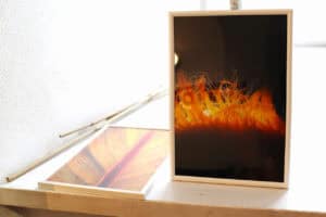 Dwa oprawione w białe ramki zdjęcia leżące na jasnym, wąskim parapecie lub półce, na tle chropowatej, białej ściany. Prawe zdjęcie stoi pionowo, przedstawiając pomarańczowe, włókniste struktury na czarnym tle. Lewe zdjęcie leży poziomo, ukazując zbliżenie na żółto-czerwony wzór liścia, z wyraźnym centralnym nerwem. Obok leżą ukośnie dwie cienkie, jasne gałązki lub patyki