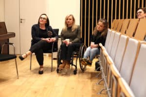 Trzy skupione kobiety. Od lewej na krześle siedzi kobieta (Anna Bieganowska-Skóra) w okularach i ciemnym żakiecie. Obok niej, centralnie, siedzi młoda kobieta na wózku (Angelika Chrapkiewicz-Gądek), trzyma kubek i patrzy w prawo. Po jej prawej stronie siedzi trzecia kobieta (Anna Dzieduszycka), która patrzy w górę i lekko na prawo. Tło tworzy jasna drewniana podłoga, rzędy pustych krzeseł z jasnymi oparciami oraz ściana z pionowymi drewnianymi panelami