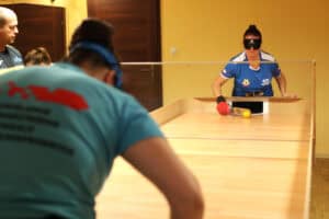 Ujęcie z meczu showdown: na pierwszym planie, tyłem do kamery, zawodniczka w jasnoniebieskiej koszulce z niebieską opaską na głowie. Po drugiej stronie drewnianego stołu, przedzielonego przezroczystą przegrodą, zawodniczka w niebieskiej koszulce i czarnej opasce na oczach, celuje rakietką (w czerwonej rękawicy) w żółtą piłkę tuż przy otworze bramkowym. Po lewej stronie w tle widoczny sędzia