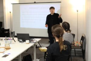 Dynamiczne ujęcie prowadzącej szkolenie (Wioletta Stępniak), która stoi przed ekranem projekcyjnym, gestykulując rękami w trakcie wykładu. Na ekranie widać kluczowy slajd z frazami do "PIERWSZEGO KONTAKTU"