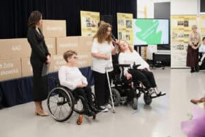 Na scenie znajduje się kilka osób. Po lewej stoi kobieta w ciemnej sukience z notatnikiem. W centrum kobieta w białej koszulce trzyma mikrofon i opiera się na lasce, przemawiając do publiczności. Po jej obu stronach siedzą na wózkach kobiety w białych koszulach, z których jedna odchyla głowę do tyłu. W tle widoczne są stosy dużych tekturowych pudeł z napisami, m.in. „AAC", „Rehabilitacja", „Wsparcie Wielka Brytania" i „Wsparcie Węgry". Po prawej znajduje się duży ekran z zieloną grafiką i napisem „Zajęcia Wspierająco-Aktywizujące", a także banery z historią stowarzyszenia