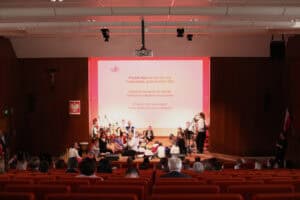 Szeroki plan sali koncertowej. Na pierwszym planie widoczna jest publiczność siedząca na pomarańczowych fotelach. Na scenie, w głębi, zespół muzyczny wraz z dziećmi (część siedzi na podłodze). Na dużym ekranie w tle wyświetlany jest tekst piosenki, której fragmenty to: "Przybyli ułani pod okienko". Po lewej stronie sceny, na ścianie, wiszą krzyż i godło Polski.